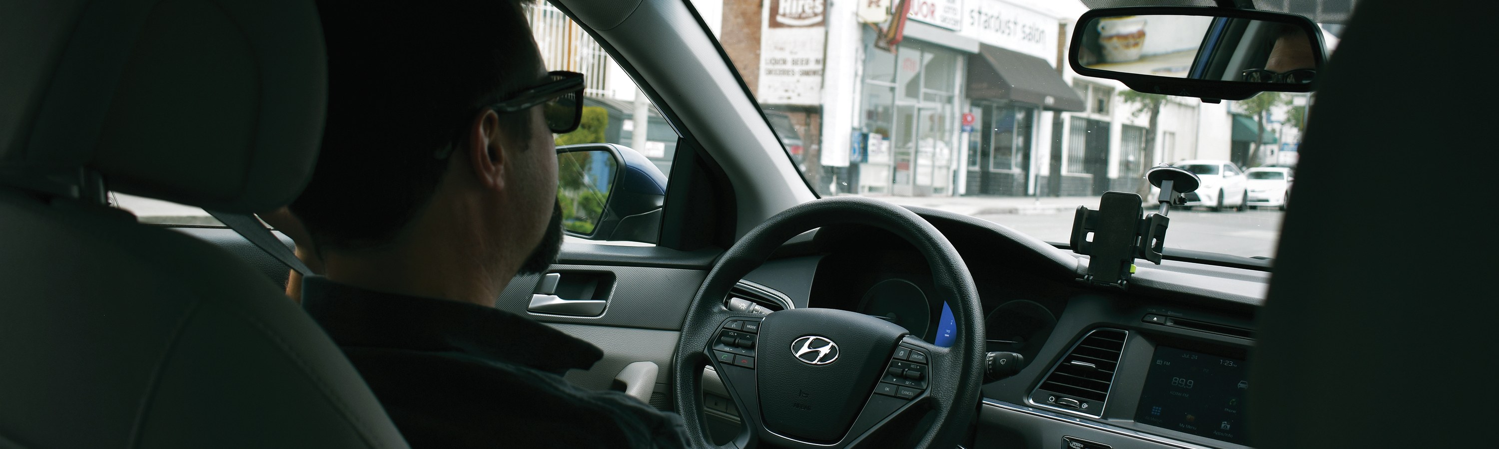 View from the back seat of a car showing a driver with hands on the wheel, a phone mounted on the dashboard, and a street of shops visible through the windshield