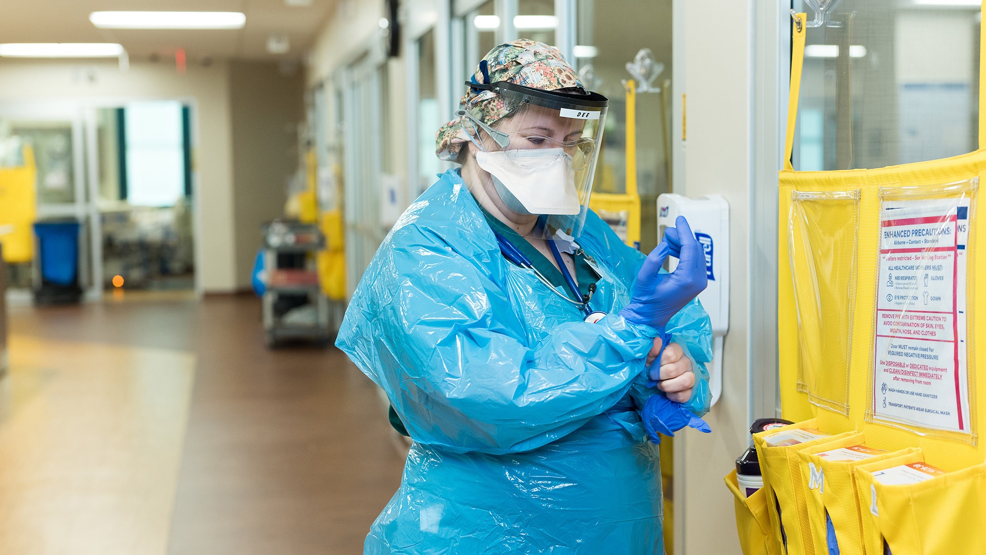 Healthcare worker in full PPE sanitizing hands beside a supply cart in a hospital hallway
