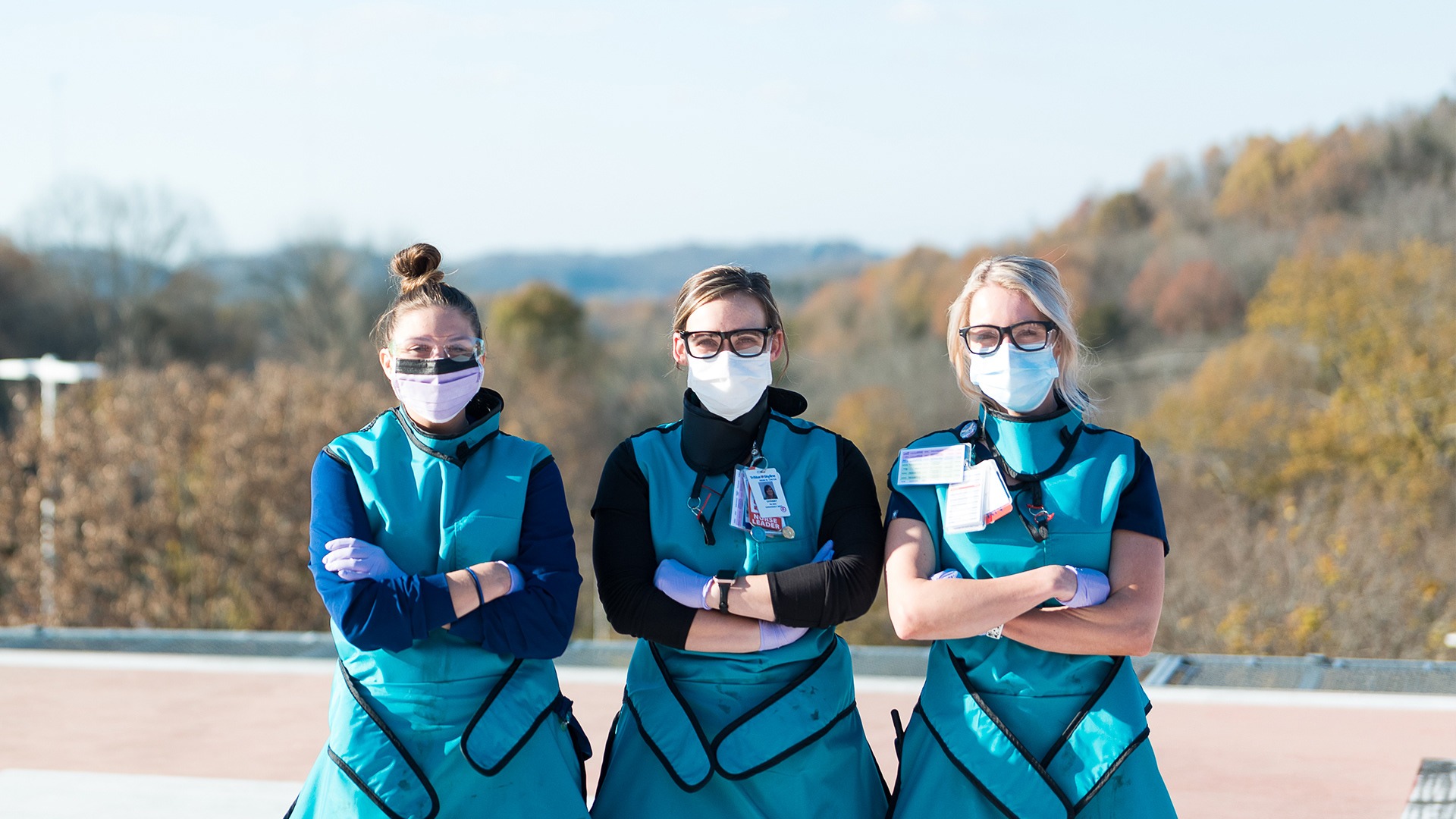 Three nurses in scrubs and masks standing outdoors with arms crossed, overlooking a wooded landscape