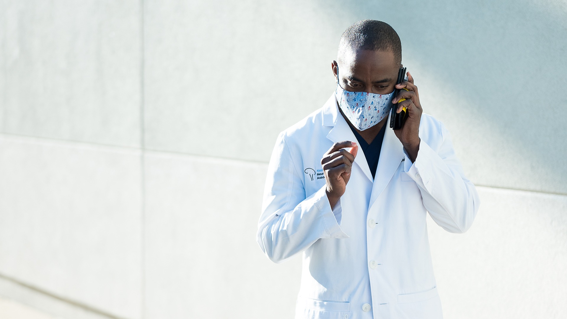 Physician in a white coat and mask speaking on the phone outside a medical building
