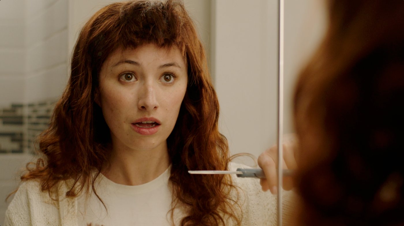 Close-up of a woman with a shocked expression holding scissors up to her hair in a bathroom mirror, from the CTV Spot commercial
